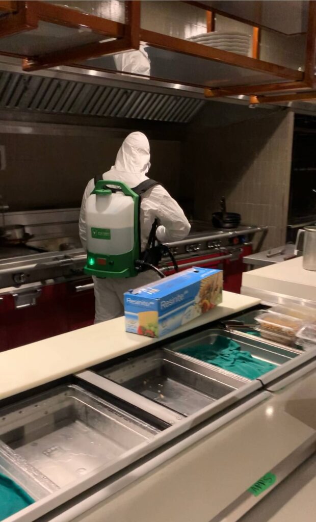 Worker in full PPE uses sanitizing sprayer under kitchen hood and on countertop equipment.