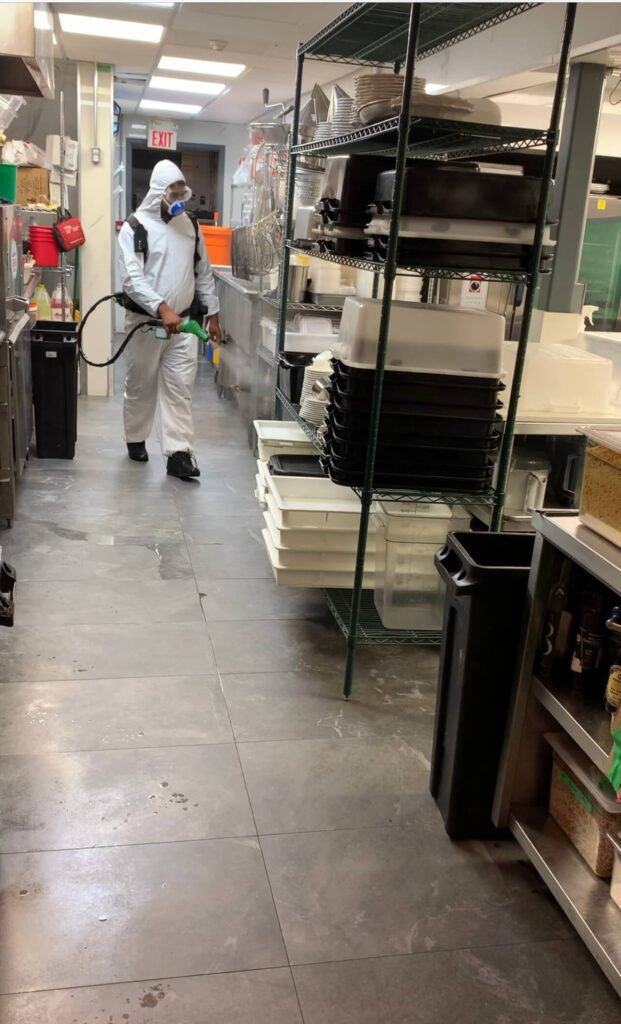 Staff in hazmat suit performing disinfection cleaning in commercial kitchen aisle with electrostatic sprayer.