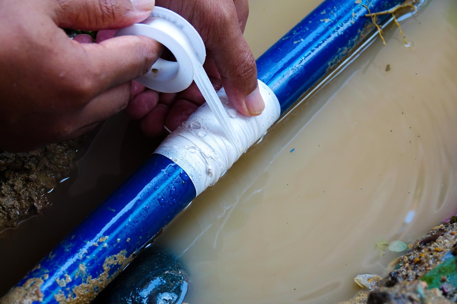 Hands sealing a leaking water pipe with plumber’s tape to stop water flow during an outdoor plumbing repair