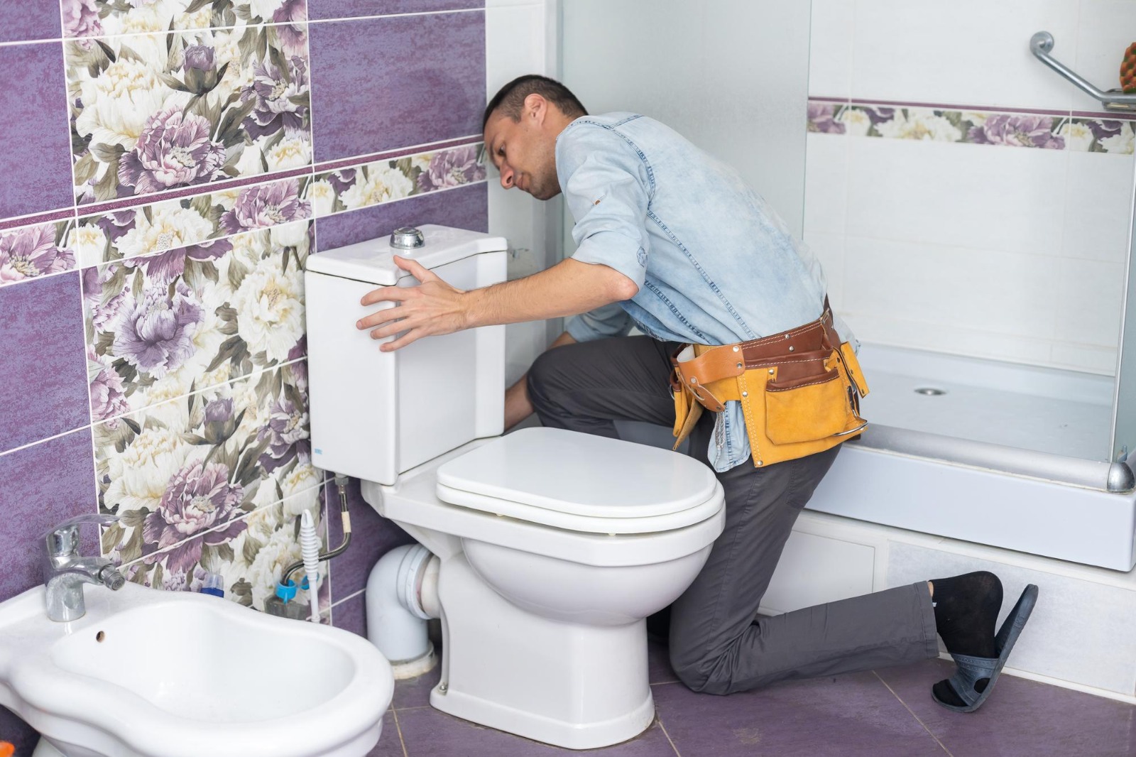 Plumber kneeling beside a toilet while inspecting and repairing bathroom plumbing.