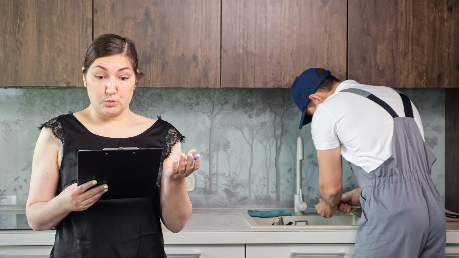 Homeowner reviewing a service checklist while a plumber repairs a kitchen sink during a home maintenance visit.